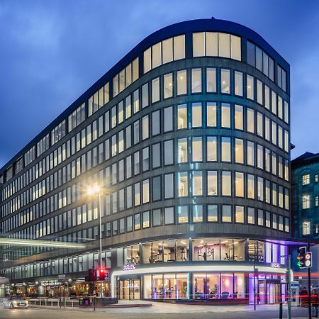 Yotel Glasgow Exterior photo The photo shows a modern office building with a distinctive curved shape and a facade made of large glass windows. It is illuminated against a twilight sky, giving it a contemporary appearance. The ground floor features large glass entrance areas, li