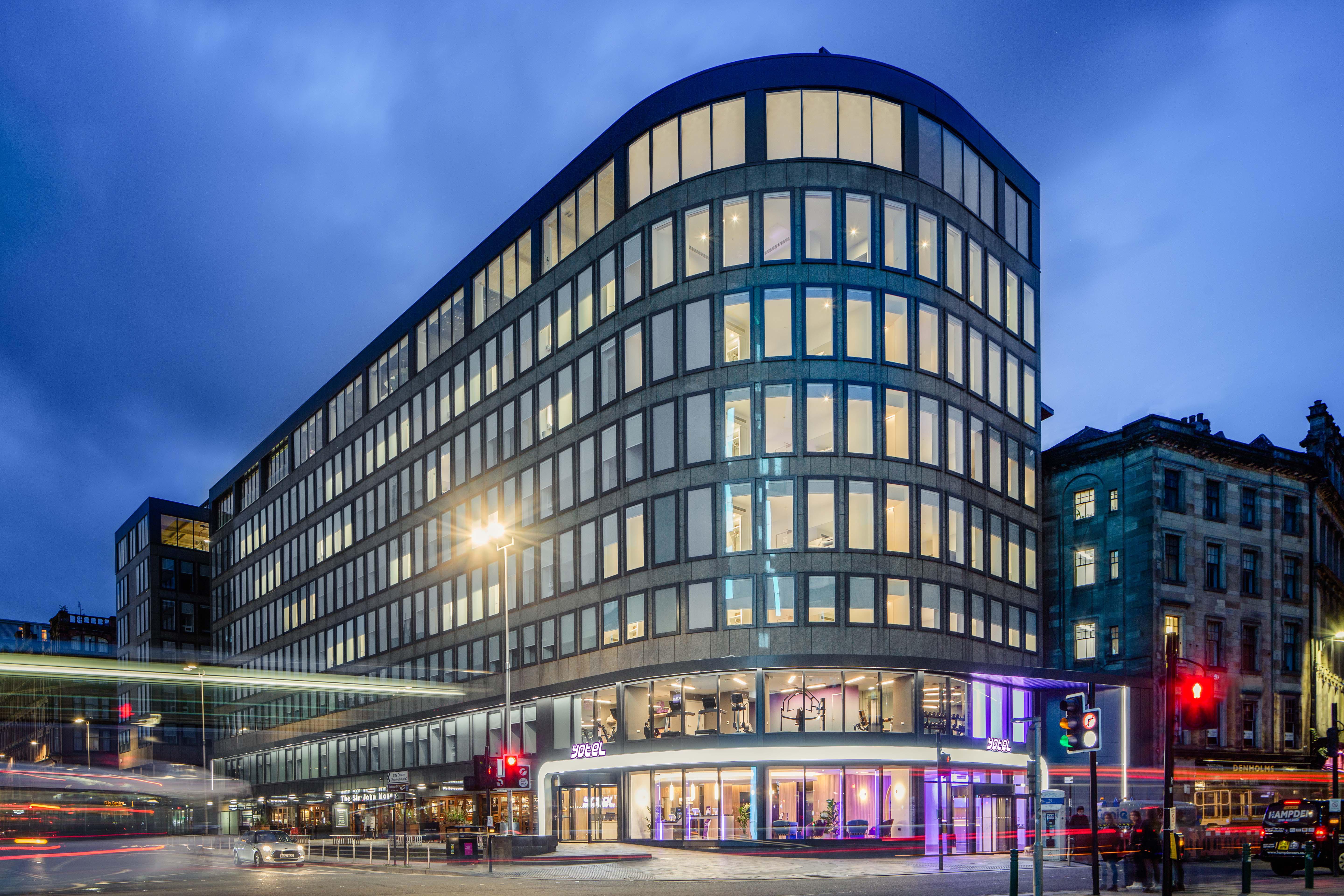 Yotel Glasgow Exterior photo The photo shows a modern office building with a distinctive curved shape and a facade made of large glass windows. It is illuminated against a twilight sky, giving it a contemporary appearance. The ground floor features large glass entrance areas, li